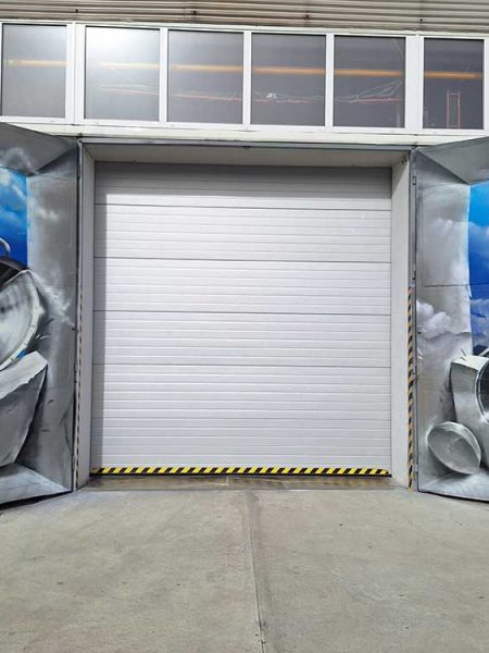Renovace starých vrat průmyslové haly výrobní firmy s mural art graffiti. Graffiti zobrazuje abstraktní kovové díly a trubky vé haly výrobní firmy s mural art graffiti. Graffiti zobrazuje abstraktní kovové díly a trubky s výrazným oranžovým akcentem, vynořující se z mraků na modré obloze.